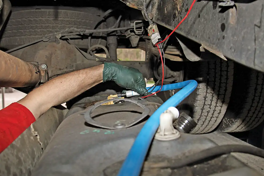 20. While lifting the fuel tank back into position he stops once it is close enough to connect the harnesses and fuel lines, including the intercept harness from the AirDog and the fuel return. Then he remounted the tank, securing it with the original straps.