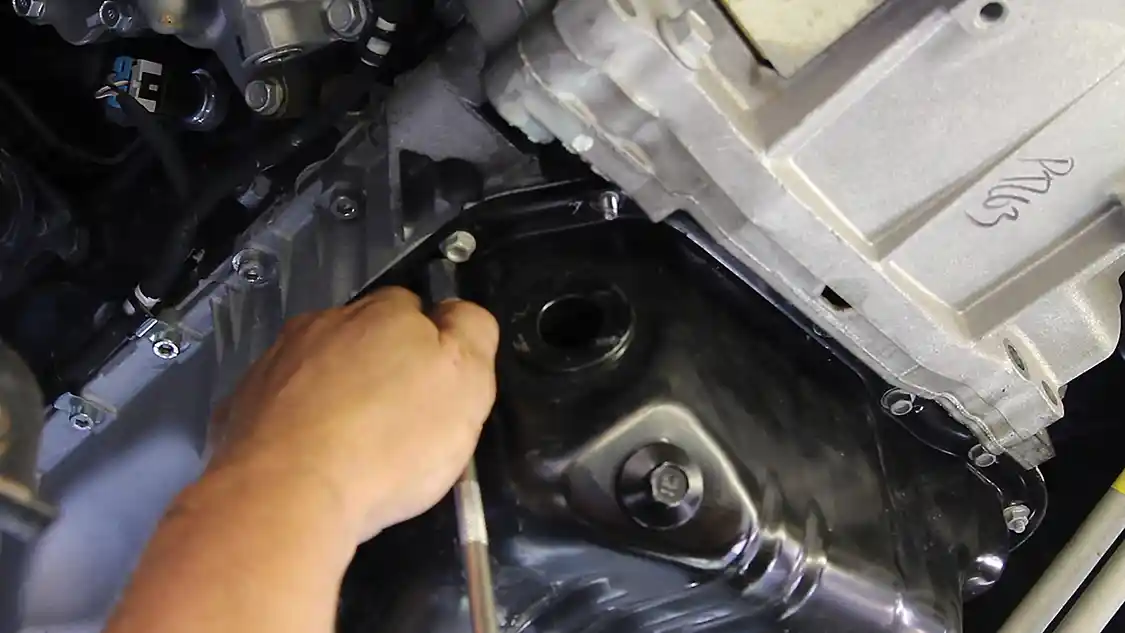 3. Use a 10mm socket with an extension to remove the two nuts and the 15 bolts holding the lower oil pan to the upper oil pan. This is the stamped sheet steel piece seen here. Note: If the transmission lines are too close, you may need to undo the retaining clip holding the lines to the engine to enable slight movement of the transmission lines. One of the bell housing bolts sticks out almost ½ in. toward the oil pan and it needs to be removed too.